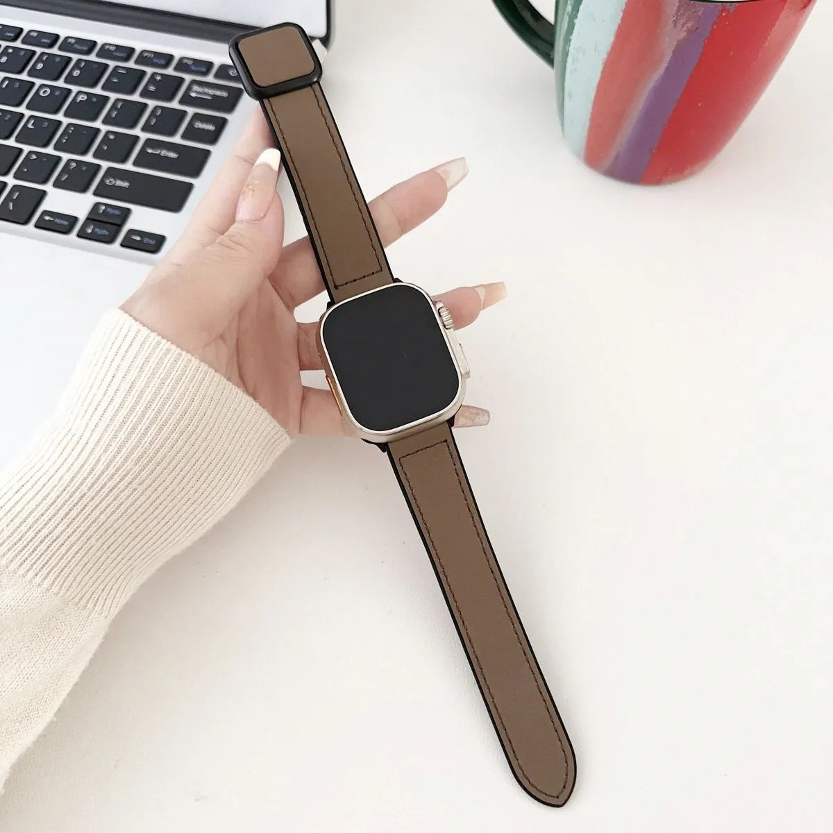 Smartwatch with brown strap held by a hand on a light surface with a laptop and mug in the background.