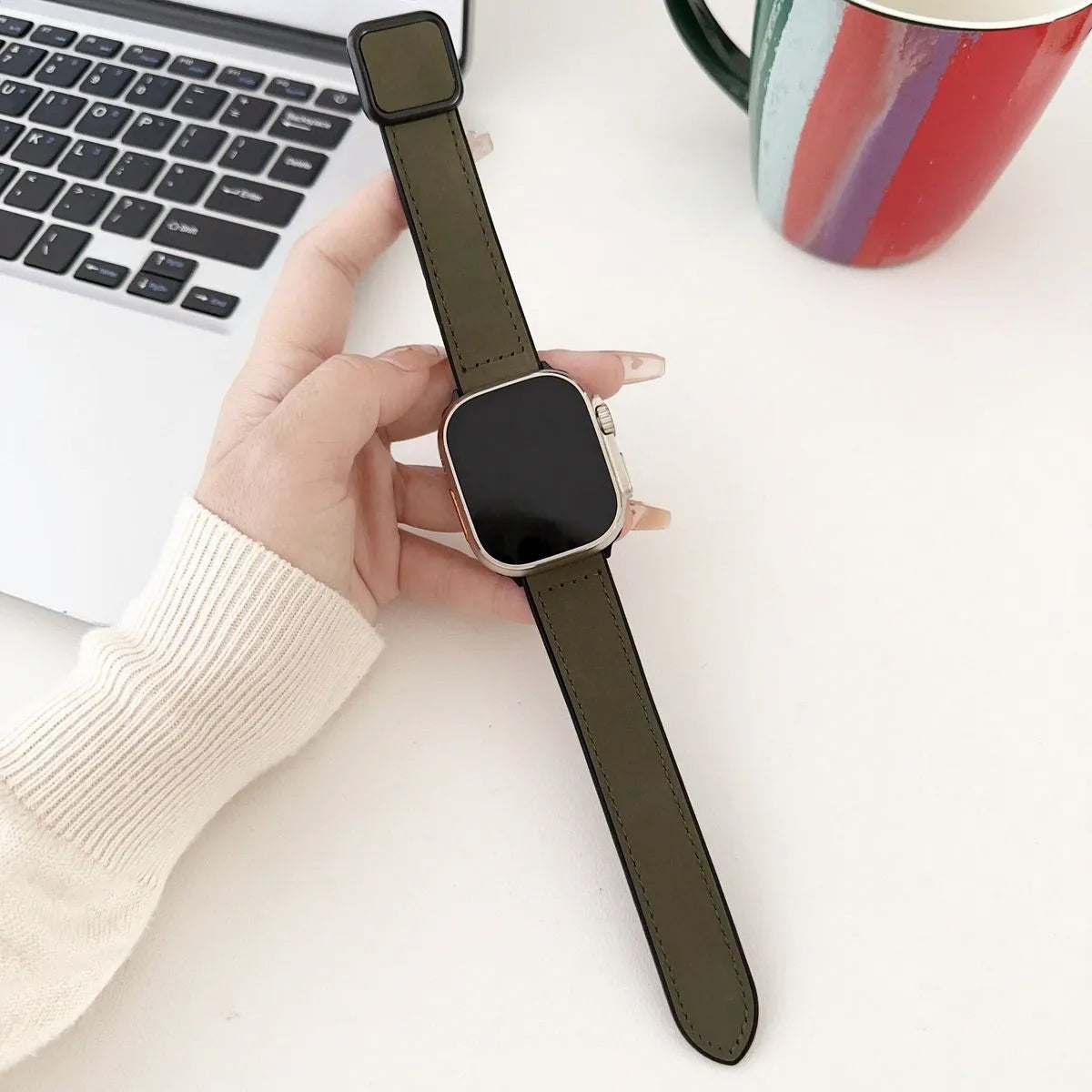 Smartwatch with a green strap held by a hand on a white surface with a laptop and colorful mug in the background.