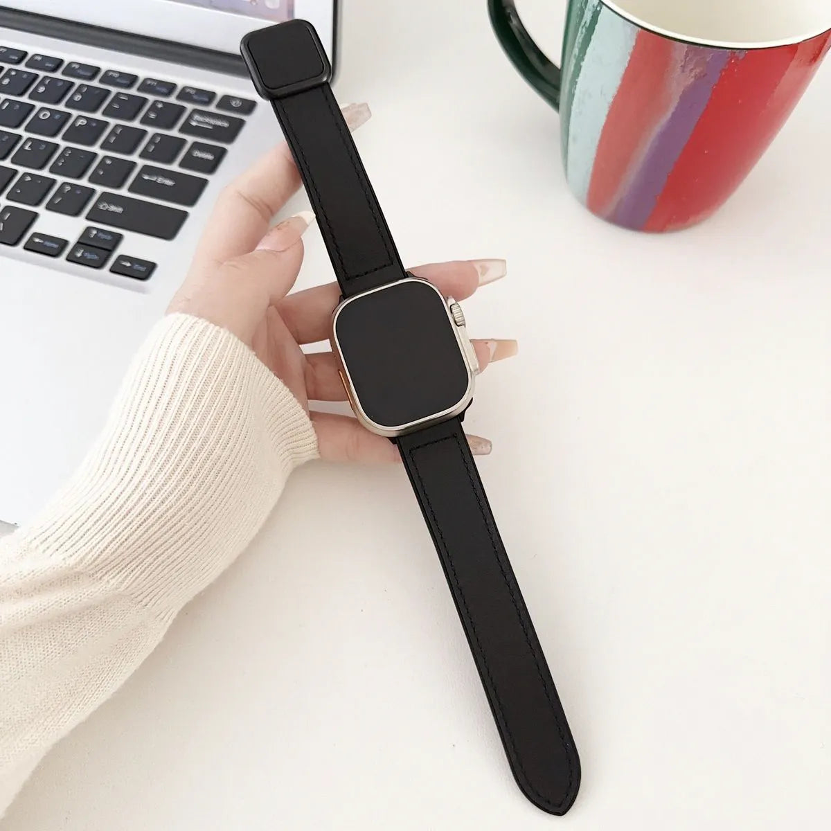Person holding a black smartwatch on a desk with a laptop and colorful mug in the background