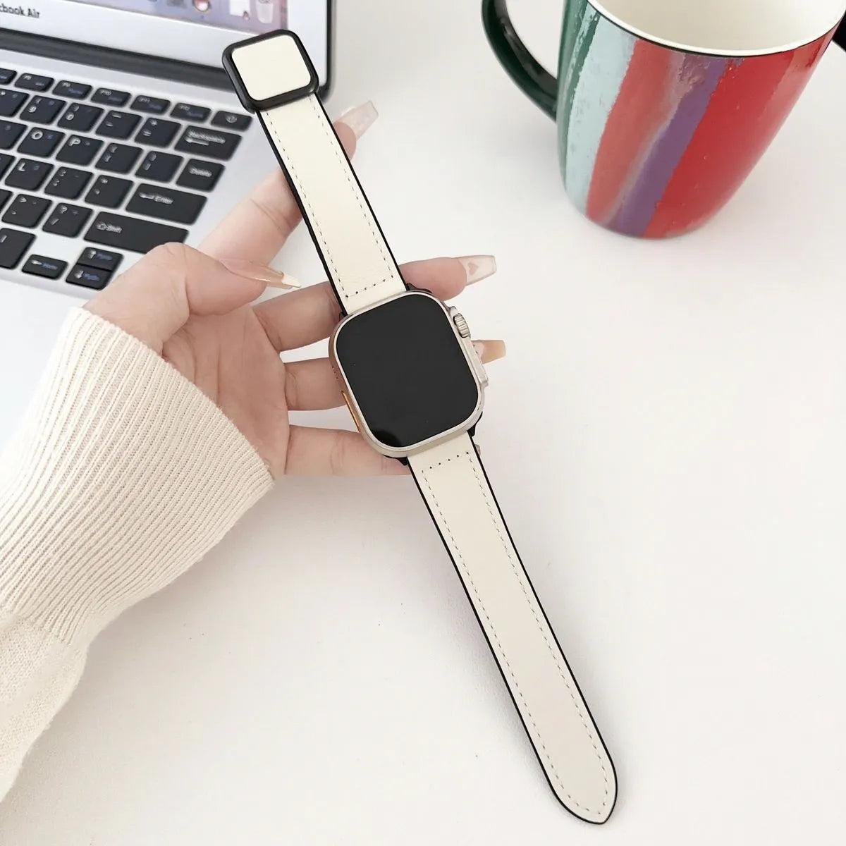 Smartwatch with a white band held by a hand on a desk with a laptop and mug in the background.