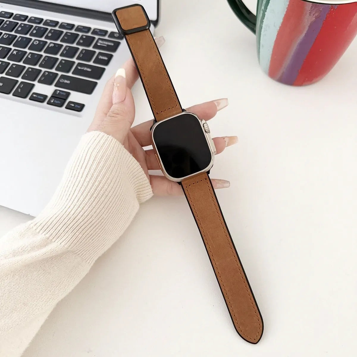Smartwatch with a brown strap held by a hand on a desk with a laptop and mug.