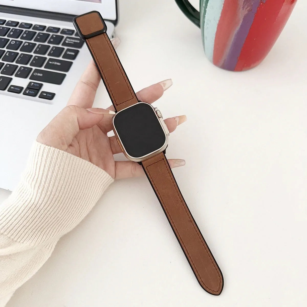 Smartwatch with brown strap held by a hand on a white surface with a laptop and mug in the background.