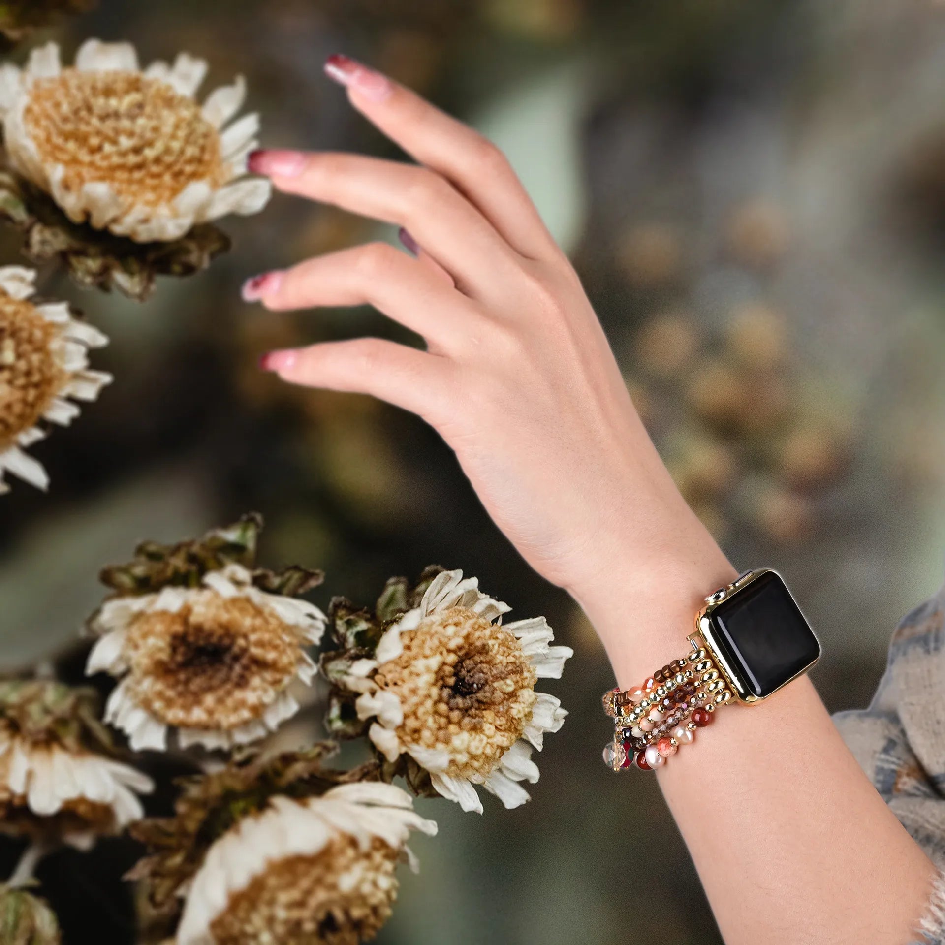 Hand wearing a smartwatch with a decorative band near dried flowers