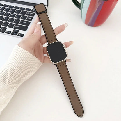 Smartwatch with brown strap held by a hand on a light surface with a laptop and mug in the background.